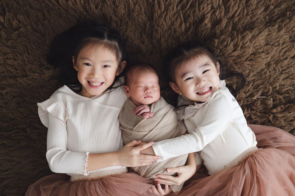 Newborn Photography Singapore Newborn photoshoot elder sisters posing with newborn baby boy on brown flokati fur rug